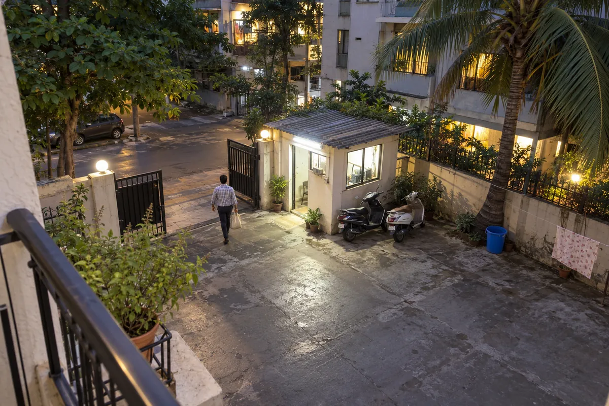 A building compound in Mumbai seen from a first-floor balcony, a man walking past the watchman cabin to the gate