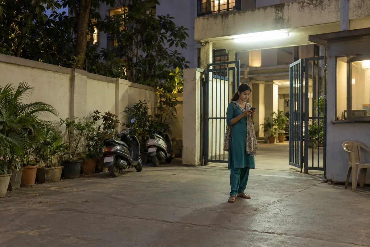 A woman in a teal salwar suit checking her phone at the gate of her building in Malad in the evening
