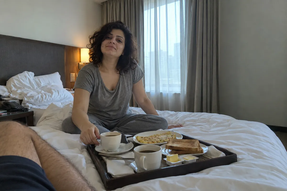 A woman in a grey t-shirt reaching for coffee from a room service breakfast tray on a hotel bed in the morning