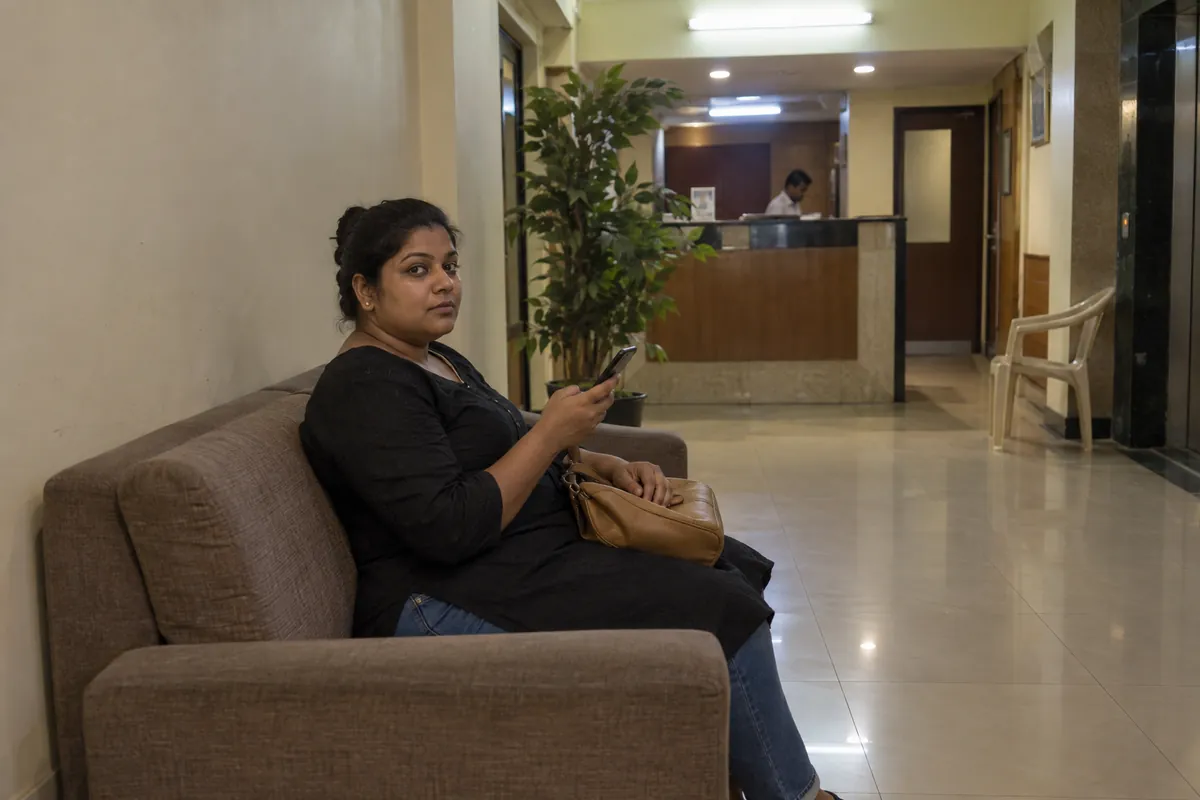 A woman in a black kurta waiting on a sofa in a small hotel lobby in Mumbai, looking up from her phone